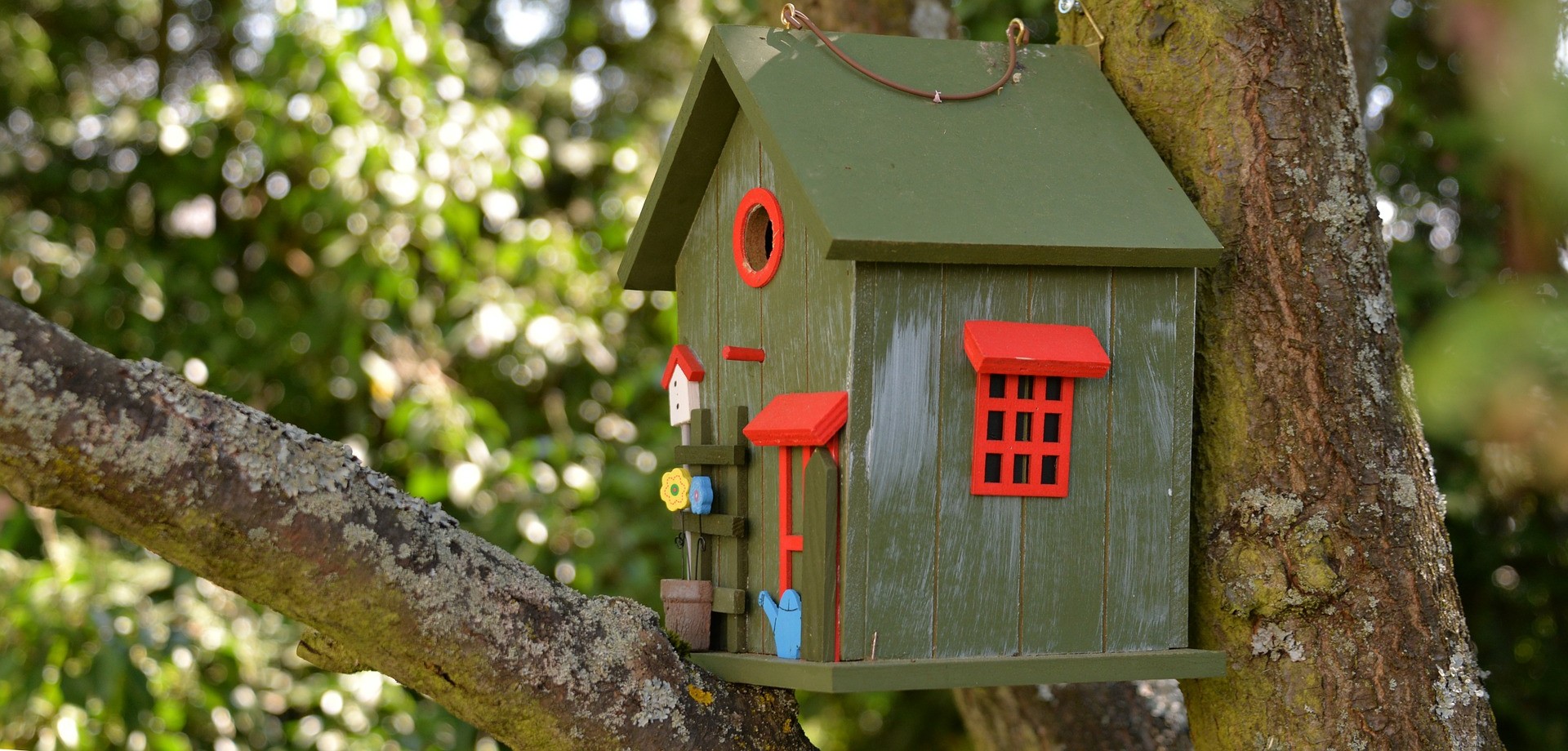 In Großaufnahme wird ein grünes Vogelhäuschen mit roten Fenstern gezeigt, davor steht eine blaue Miniatur-Gießkanne und kleine Miniatur-Blumen