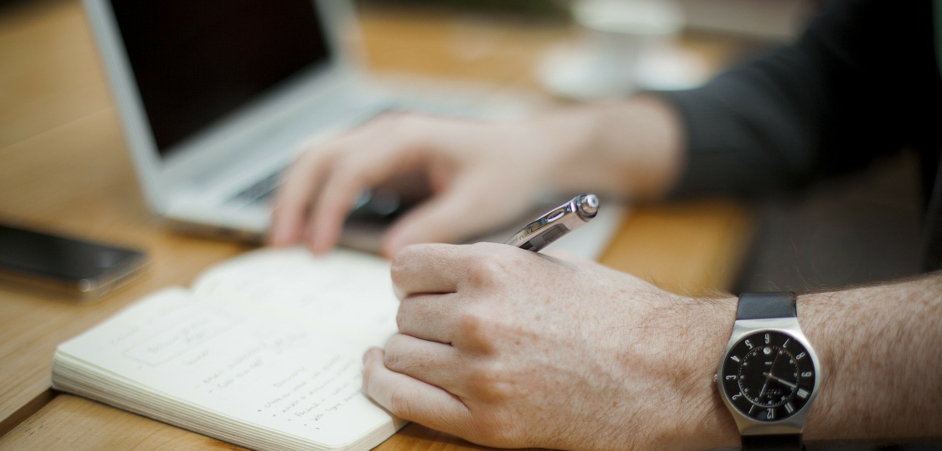Ein Mann sitzt an einem Schreibtisch und notiert sich Gedanken mit einem Kugelschreiber in ein Notizbuch, daneben steht ein aufgeklapptes Notebook